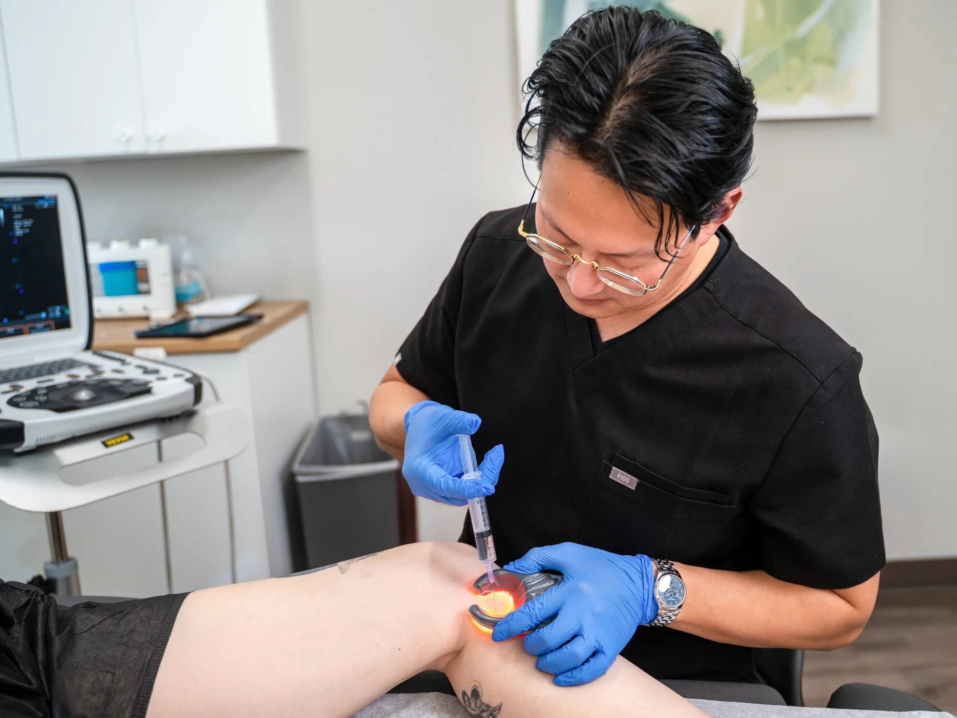 Dr. Benjamin Kum performs a sclerotherapy procedure on a patient's leg, using a Veinlite to illuminate the target vein while injecting a sclerosant with a syringe. An ultrasound machine is visible in the background, assisting with the clinical assessment.