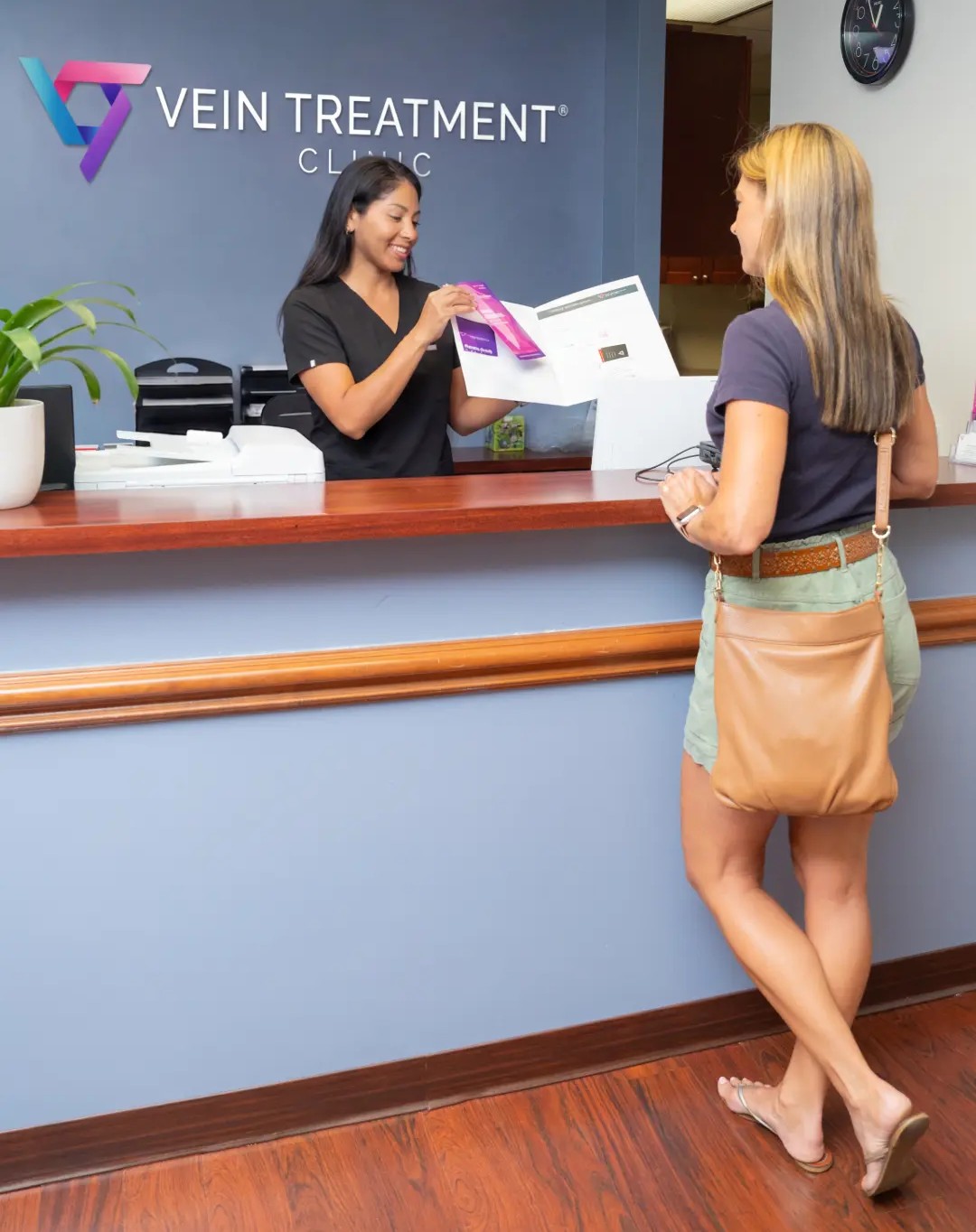 Frontdesk A receptionist smiles as she hands a "Pre-Procedure Instructions" document over a front desk to a female patient. In the background, a blue wall features a white sign reading "VEIN TREATMENT CLINIC," and a round black clock hangs on an adjacent white wall.
